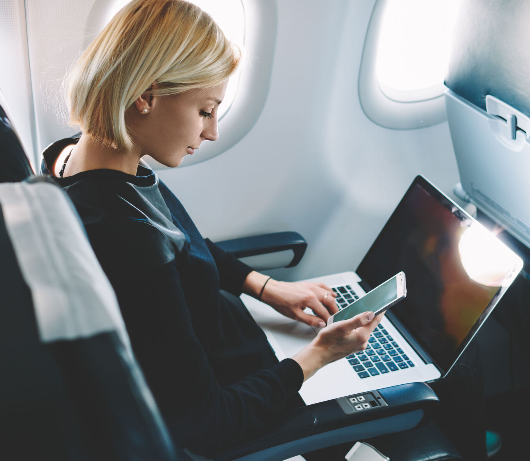 Businesswoman with short haircut sitting in airplane cabin and chatting online on smartphone while checking email on laptop computer with mock up area.Female traveler reading notification on cellular